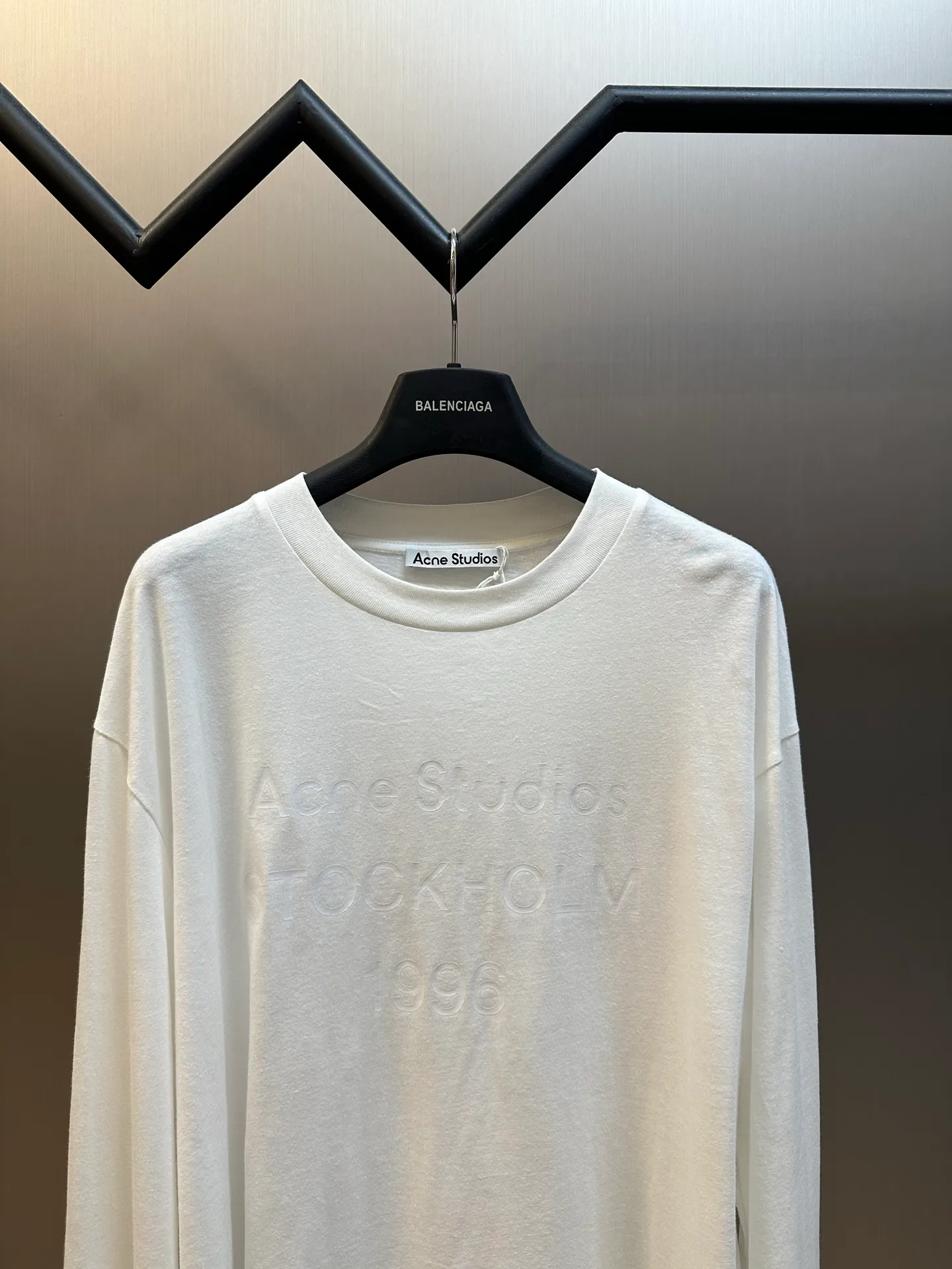 Ac Original White Letter Round Neck Long Sleeve The fabric is made of 32-count cotton and linen blended plain knitting, with a ratio of 85% cotton and 15% linen fiber. The weight is only 145g/m²; the ribbed neckline is made of Same Style cotton and linen blended yarn, 145g/m², to ensure a unified texture of the entire piece. The collision of the natural rough base and the industrial spraying process creates a natural irregular texture and an extremely light and breathable wearing experience. Cotton provides a skin-friendly and soft foundation, while linen introduces natural roughness and knotted particles, giving the fabric surface an irregular texture. The addition of hemp fiber makes the fabric have excellent moisture absorption and perspiration properties, while also bringing a natural micro-wrinkle texture and dry touch. After the fabric is piece-dyed, the dye evenly penetrates into the fiber, showing full color fastness and soft matte color, providing a uniform base for subsequent inkjet printing. The printing uses a double-layer printing process of environmentally friendly elastic glue and varnish, which takes into account the concept of environmental protection, wearing comfort and visual expression. The bottom layer uses environmentally friendly elastic glue, with water-based environmentally friendly resin as the base material. The color paste is scraped onto the surface of the fabric through a high-mesh screen. After drying and solidifying at high temperature, a soft and highly elastic continuous adhesive film is formed. The printed film can stretch and retract with the fabric. Even if it is covered with a large area of ​​pattern, it is still breathable and does not feel tight when wearing it. The surface layer is accurately overprinted with transparent varnish, creating a high-gloss effect in specific areas. This layer of varnish not only significantly improves the color contrast of the pattern, but also creates a local wet and bright visual and tactile contrast, making ordinary printing instantly gain three-dimensional layers and glossy texture. At the same time, the smooth surface layer also enhances Wear-Resistant and wash-resistant properties. This T-shirt is defined as a casual loose cut, unisex style, and hip length. The light fabric slides smoothly along the body contours, forming natural and soft folds at the shoulders and waist. The shoulder line falls naturally, the bust and hem circumferences are similar, and it presents a straight silhouette, without deliberately distinguishing between male and female body shapes. The hip-length design utilizes the elongated body and the extremely light drape of the fabric to create lazy and flowing lines. The ribbed neckline, cuffs and hem are all made of Same Style cotton and linen blended yarn, which maintains a unified overall texture and is loose yet exquisite in details. size：xs s m l
